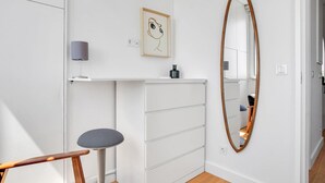 A compact, modern room with a white dresser, a round mirror, a framed artwork, and a small stool.