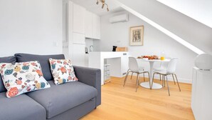 A modern apartment with a grey sofa, white dining table, and a kitchen area.