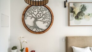 Una habitación con un cuadro redondo de un árbol, una cama con cabecera color beige, una mesita con un jarrón, una botella y un cuenco con fruta.