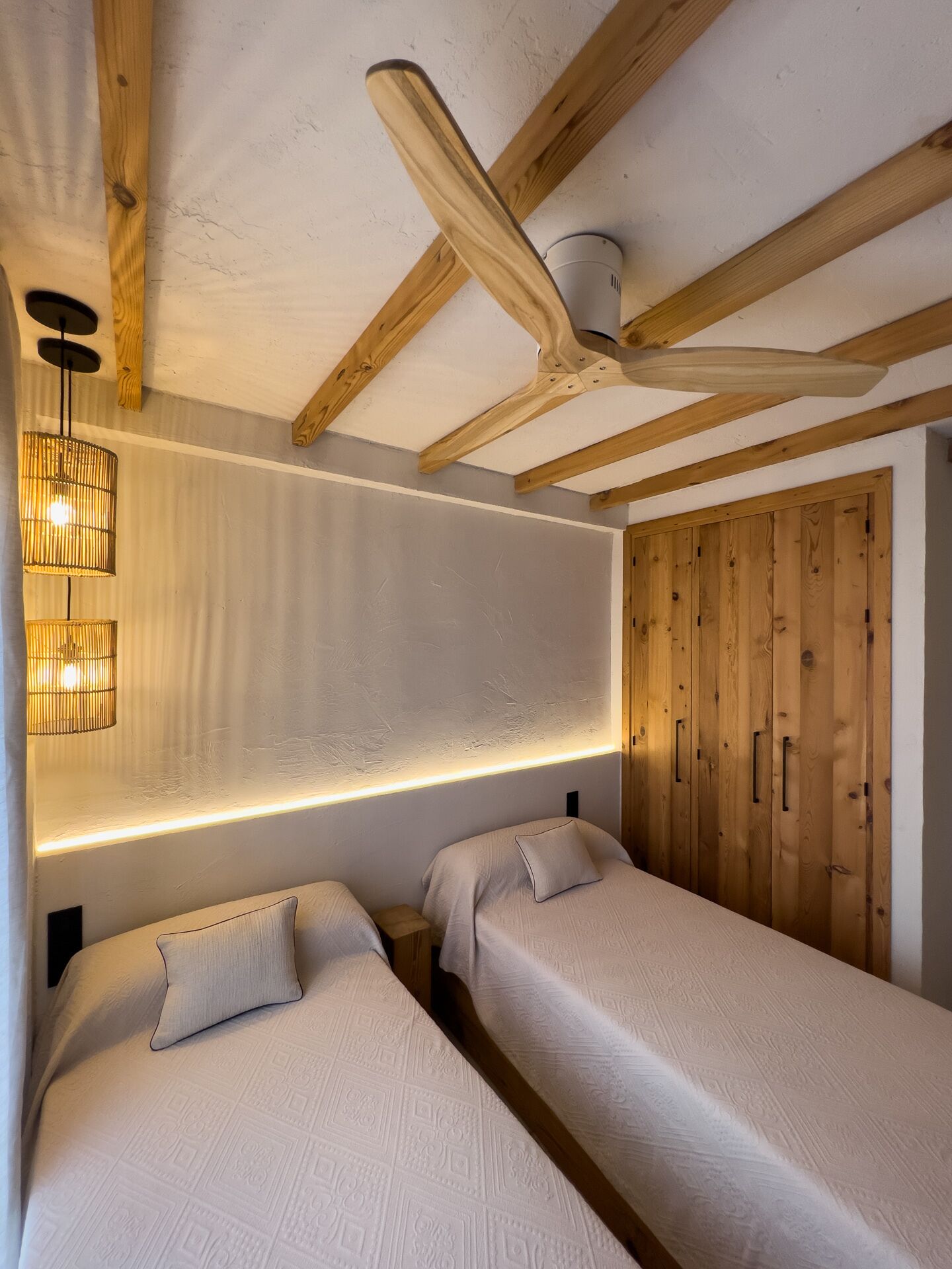 A room with two beds, wooden ceiling beams, a ceiling fan, and a wall-mounted light fixture.