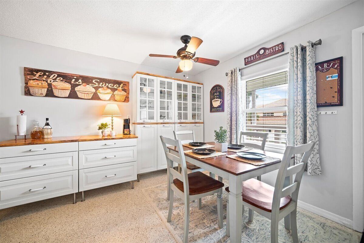 A dining area with a wooden table and white chairs, a ceiling fan, and a window with curtains.