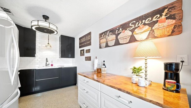 Een moderne keuken met zwarte kasten, witte aanrechtbladen en een houten kookeiland. Er staat een koffiezetapparaat, een lamp en een bordje met de tekst 'Life is Sweet' en afbeeldingen van cupcakes.