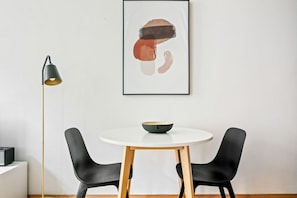 A minimalist room with a round white table, two black chairs, a black floor lamp, and a framed abstract artwork on the wall.