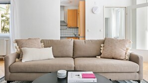 Una sala de estar moderna con un sofá beige, dos almohadas, una mesita de centro con un libro y una cocina al fondo.