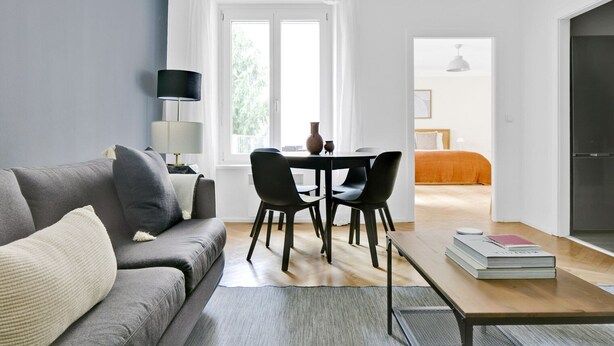 Una sala de estar moderna con un sofá gris, una mesa de centro y un comedor con sillas. Hay un ventanal con cortinas, un televisor de pantalla plana y una estantería.