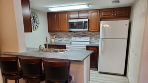 A kitchen with wooden cabinets, a stainless steel sink, a microwave, a stove, a refrigerator, and a bar area with stools.