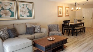 A living room with a beige sofa, patterned cushions, a wooden coffee table, and a dining area with a wooden table and chairs.