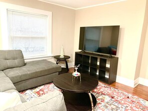 Una sala de estar con un sofá modular, una mesa de centro redonda con un objeto decorativo, un televisor sobre un mueble bajo y una alfombra estampada.