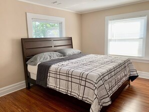 A bedroom with a bed, wooden headboard, and two windows with blinds.