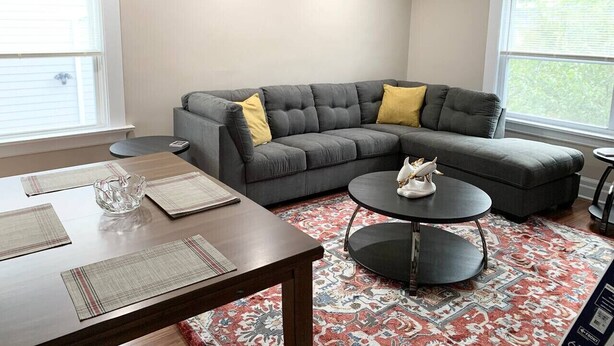 A living room with a gray sectional sofa, a round coffee table, and a dining table with placemats.