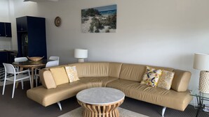A modern living room with a beige sectional sofa, a round coffee table, and a dining area with a table and chairs. - Sea Eagles 10 Steps To Kings & Shelly Beach (KINGS BEACH)