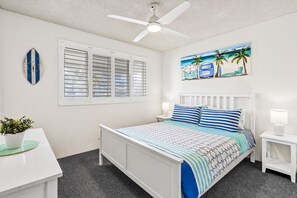 A bedroom with a bed, a ceiling fan, a wall clock, a window with blinds, a painting, and a bedside table. - Edmund Shores 6 Walk To Kings Beach, Ocean Views (KINGS BEACH)