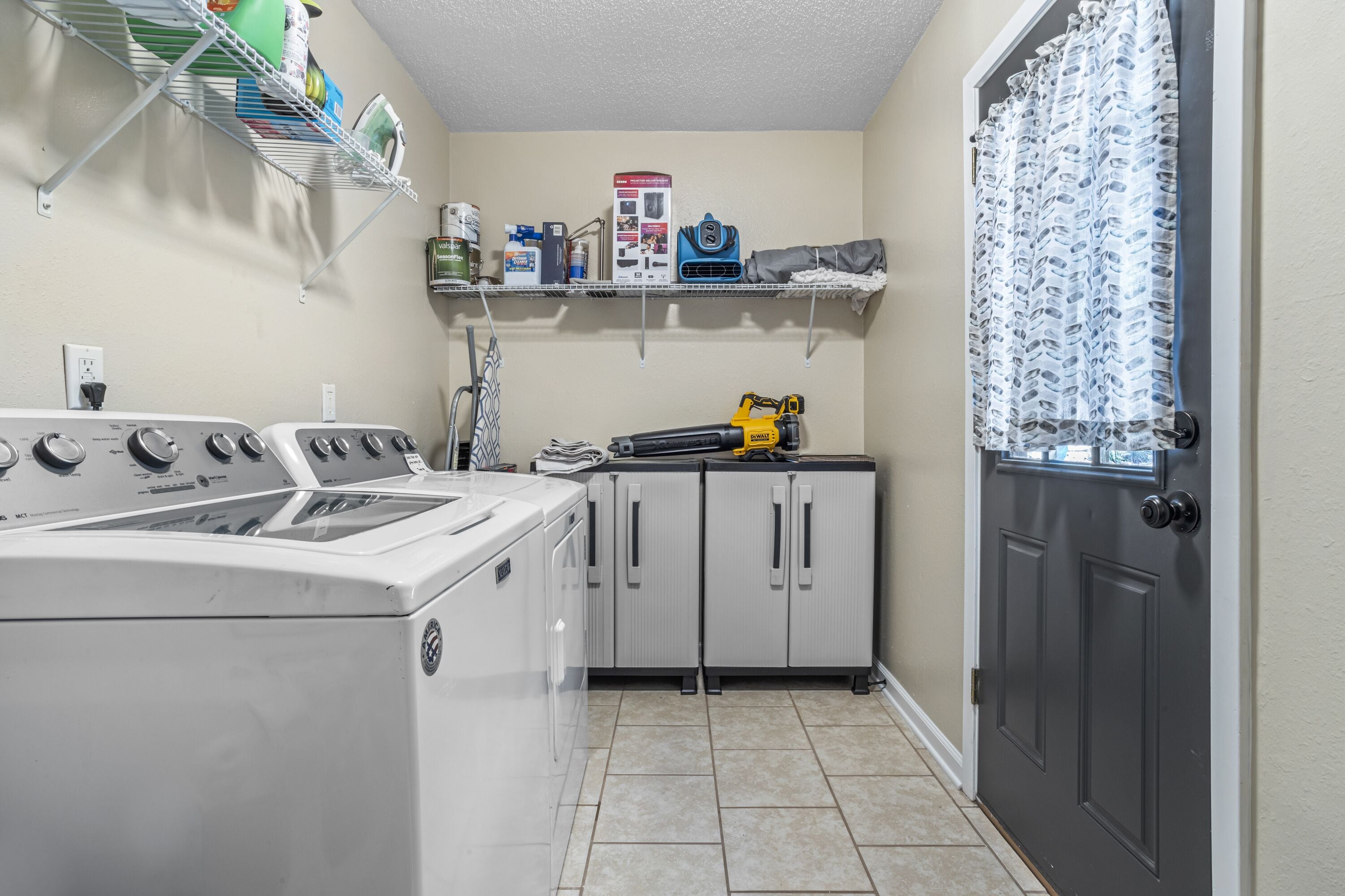A laundry room with a front-loading washer and dryer, a metal shelf with cleaning supplies, and a gray door with a patterned curtain.