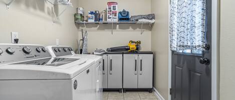 A laundry room with a front-loading washer and dryer, a metal shelf with cleaning supplies, and a gray door with a patterned curtain.
