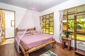 Una habitación con cama, cabecera de madera, mosquitera, ventana con cortinas y una mesita con ventilador.