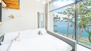 A bedroom with a large window overlooking a coastal landscape, a bed with white bedding, and a small shelf with decorative items. - Yeosu Sea&blue Family Pension (Yeosu)