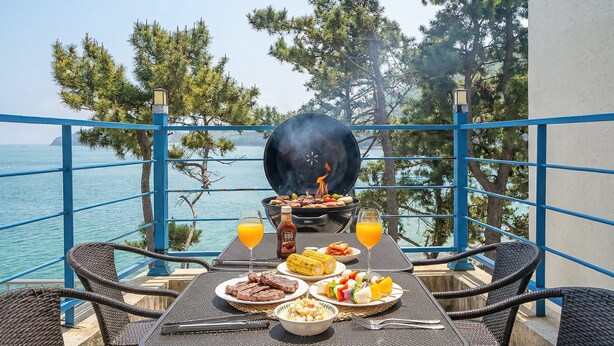 A balcony with a grill, table with plates of food, drinks, and a view of the sea. - Yeosu Sea&blue Family Pension (Yeosu)