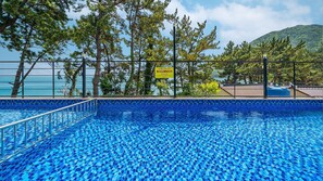 A clear blue pool with a tiled edge, surrounded by a metal fence, with a view of the sea and trees in the background. - Yeosu Sea&blue Family Pension (Yeosu)