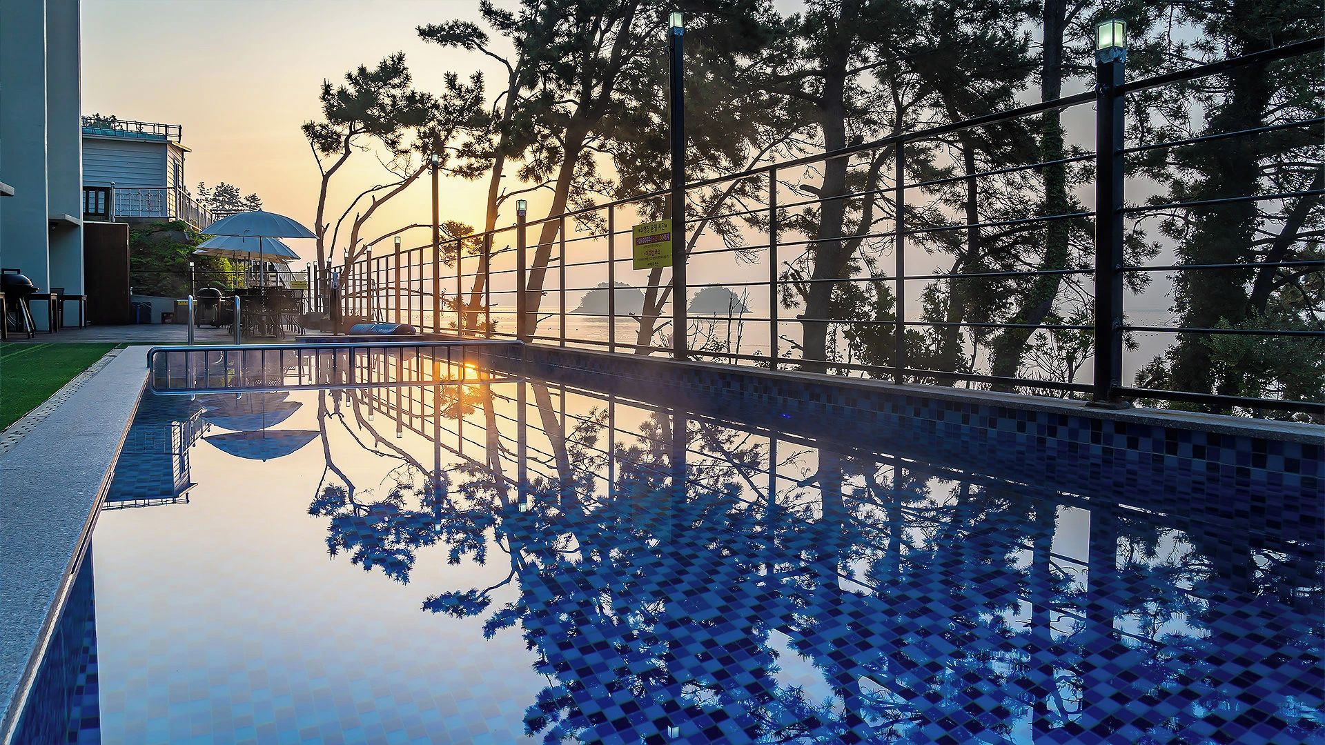 A swimming pool with a tiled deck, surrounded by trees and a fence.