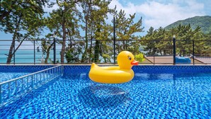 Ein Schwimmbecken mit einer gelben Luftschwan, umgeben von einem Metallzaun, mit Bäumen und Blick auf das Meer im Hintergrund.