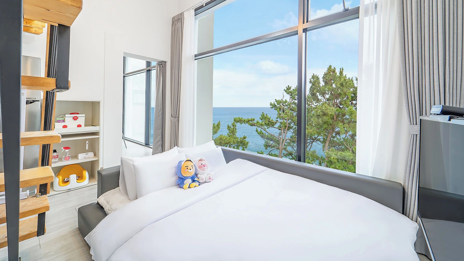 A hotel room with a large bed, a view of the sea, a desk with a computer, and a shelf with toiletries.