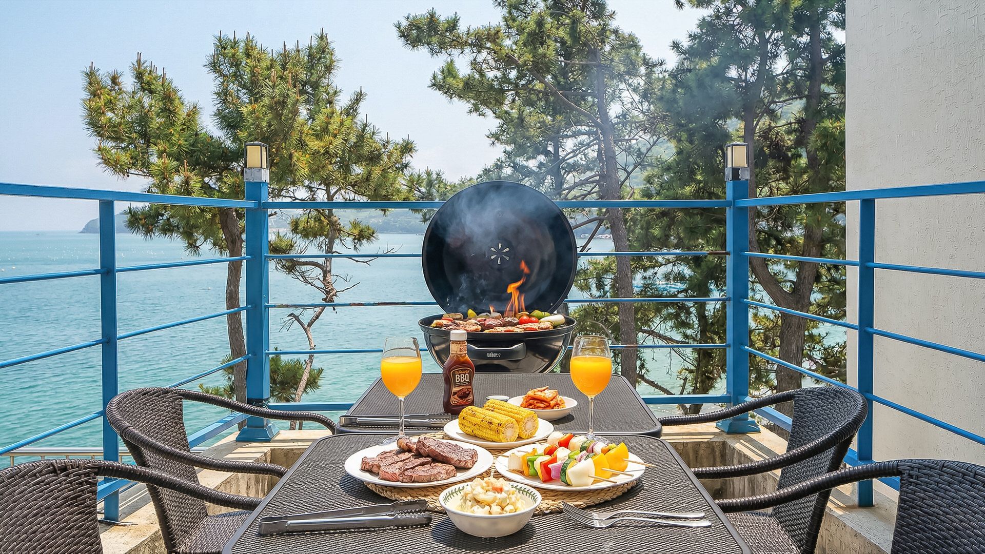 A balcony with a grill, table with food, drinks, and a view of the sea.