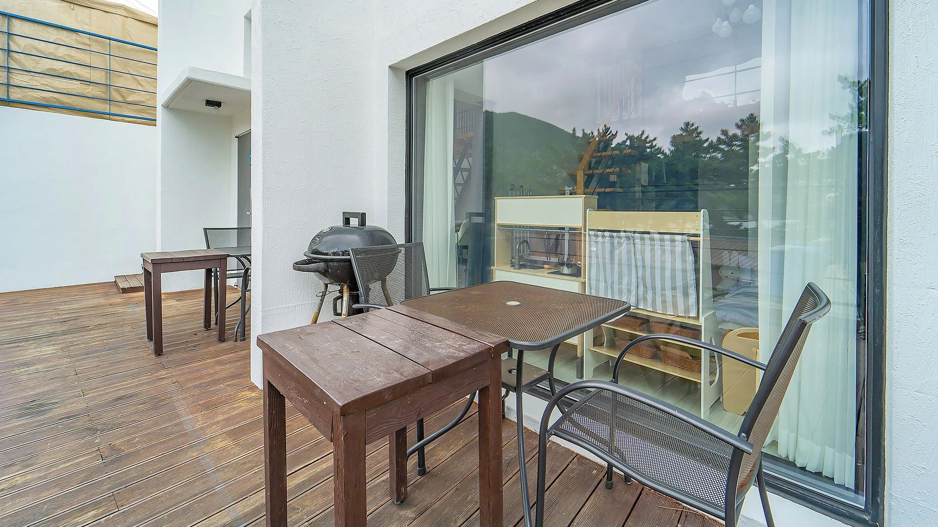 A wooden deck with a table and chairs, a grill, and a view of a garden through a large window.
