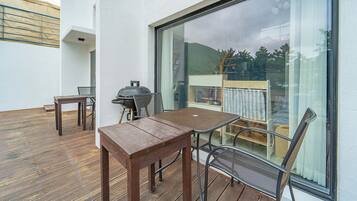 A wooden deck with a table and chairs, a grill, and a view of a garden through a large window.