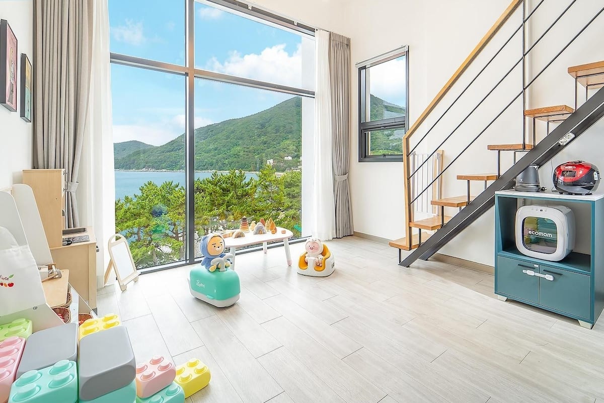 A spacious room with a large window offering a view of a lake and hills, a wooden staircase, and a teal cabinet with a vintage television.