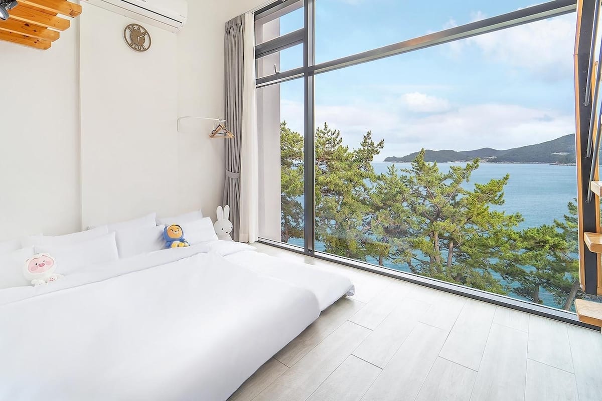A bedroom with a large window offering a view of a body of water and trees, a bed with white bedding, a clock on the wall, and a hanging coat rack.