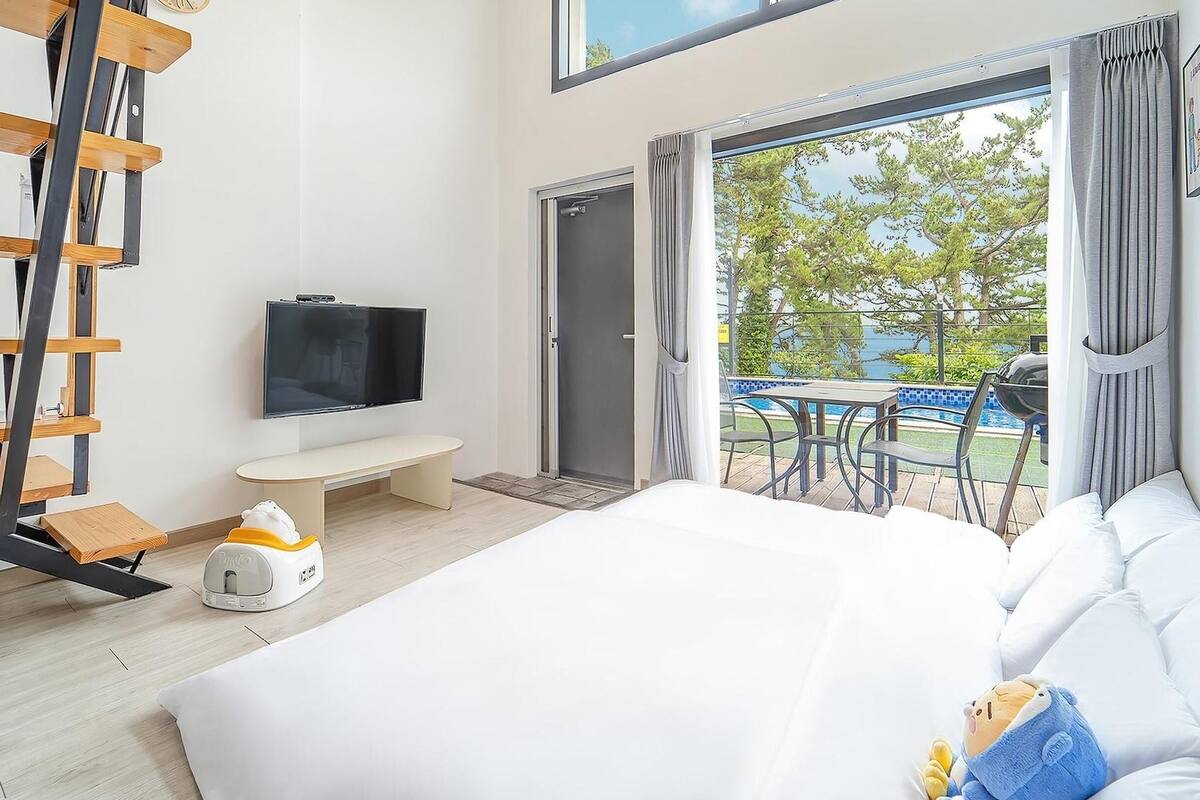 A modern hotel room with a bed, a TV, a bench, a small table, and a balcony with a pool view.
