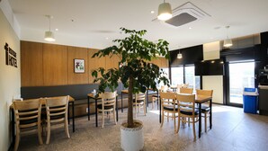 A hotel lobby with wooden panel walls, a central potted tree, multiple wooden tables and chairs, and large windows allowing natural light. - Chungjangro 1st Boutique Hotel (Gwangju)