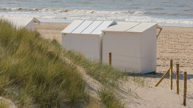 Une plage avec deux cabanes de plage blanches, un rivage sablonneux et un voilier à l’horizon.