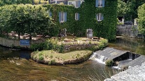 Un edificio lungo un fiume con un ponte in pietra, una piccola cascata e vegetazione lussureggiante.
