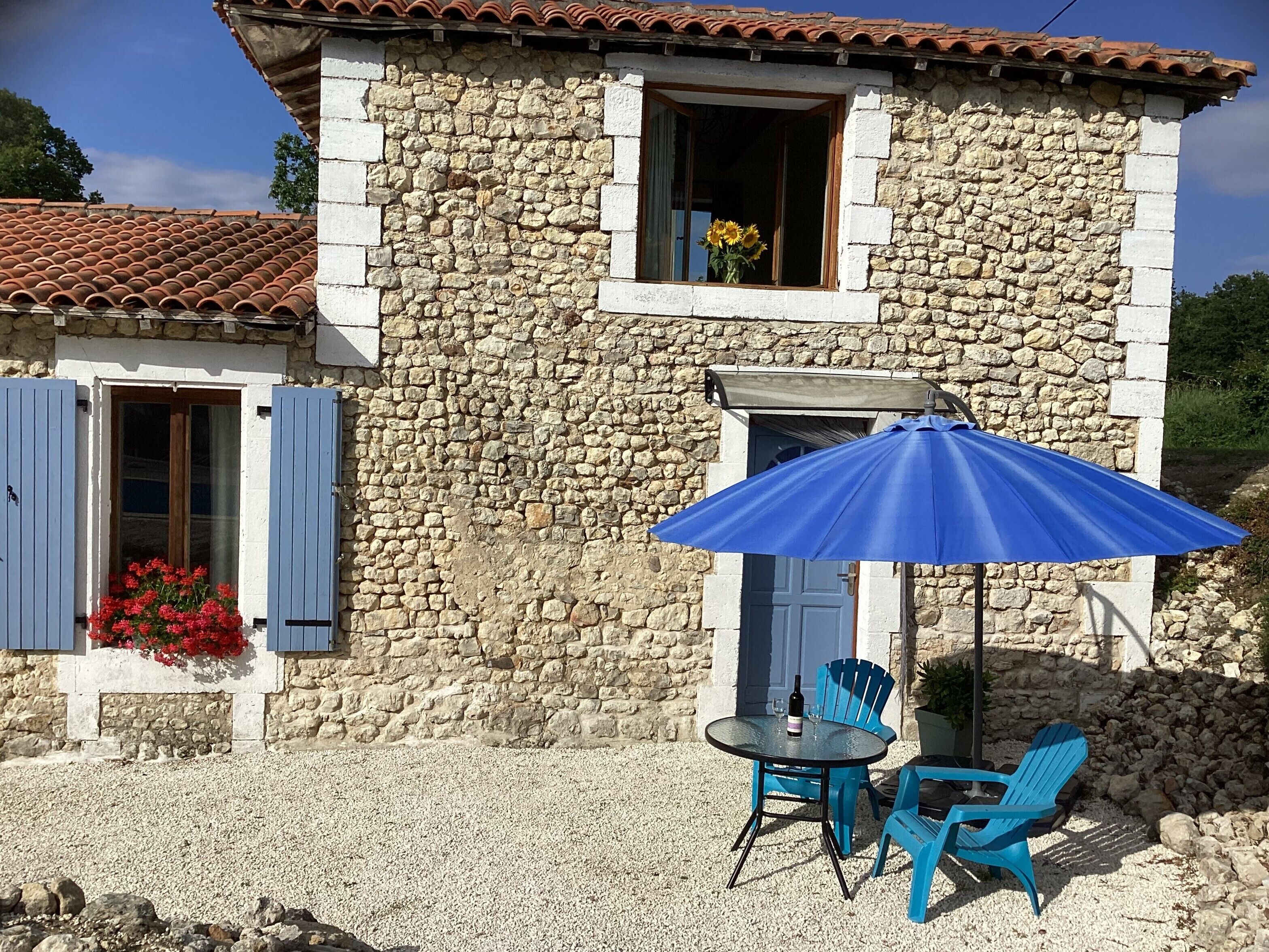 Un bâtiment en pierre avec un toit en tuiles, une fenêtre avec un volet bleu et un patio comprenant une table et des chaises.