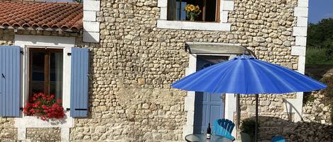 A stone building with a tiled roof, a window with a blue shutter, and a patio area with a table and chairs.