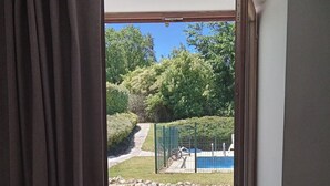 A view through a window with a curtain, revealing a garden with a pathway, a pool, and a stone wall. - Chez Goret Gites - Gite Skippy  Couples only (Nouvelle- Aquitaine)