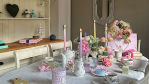 Mesa de jantar posta com xícaras de chá, um bule e diversos objetos decorativos, incluindo flores e um espelho.