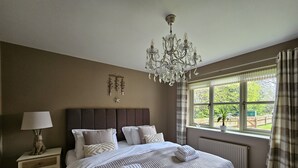 Quarto com uma cama grande, mesa de cabeceira com luminária, um lustre e janela com cortinas.