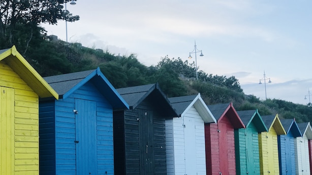 Uma fileira de cabanas de praia coloridas com portas numeradas, com árvores e um céu nublado ao fundo.