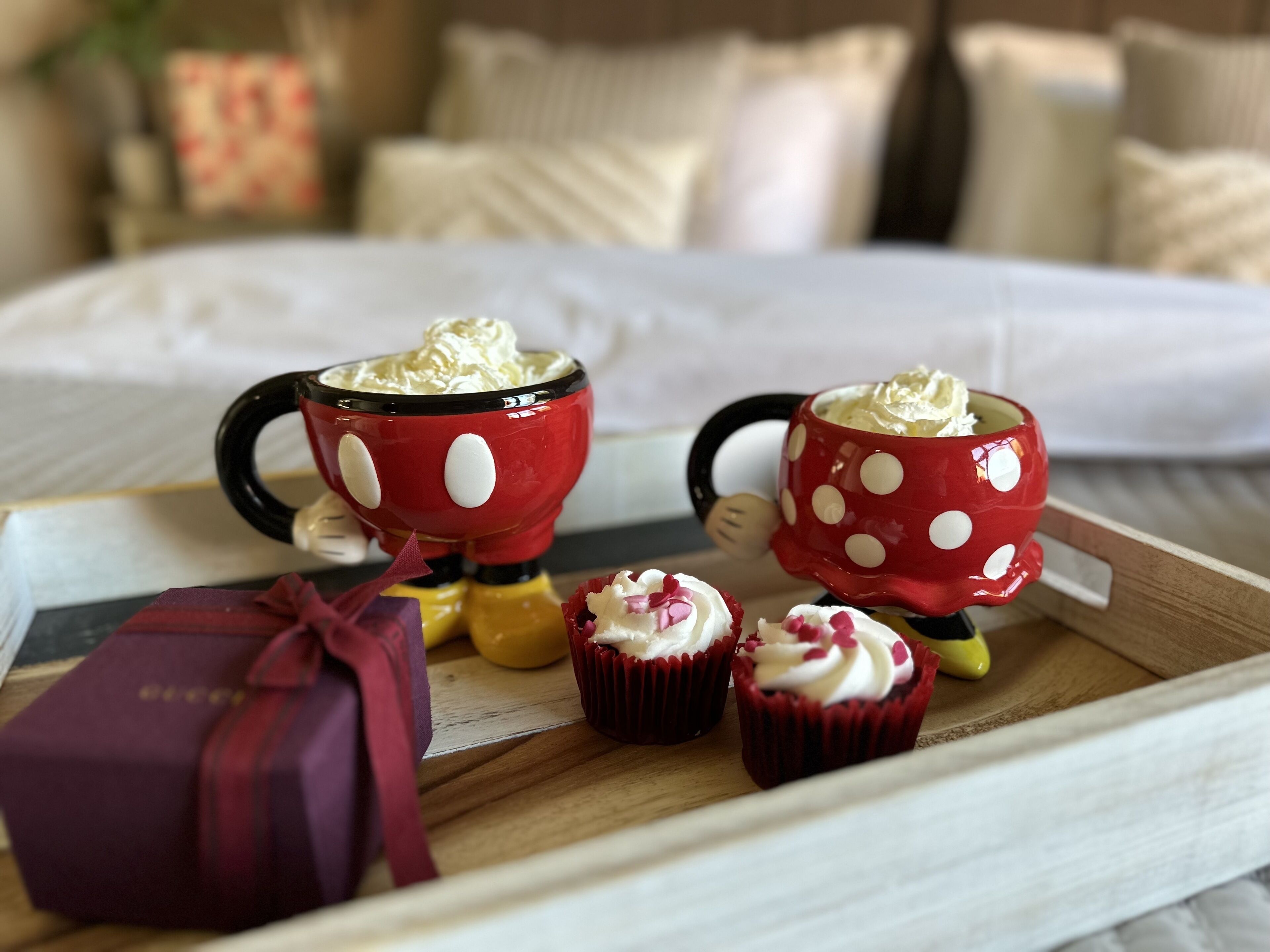 A tray with two Mickey Mouse mugs filled with whipped cream, a gift box, and two cupcakes on a wooden surface.