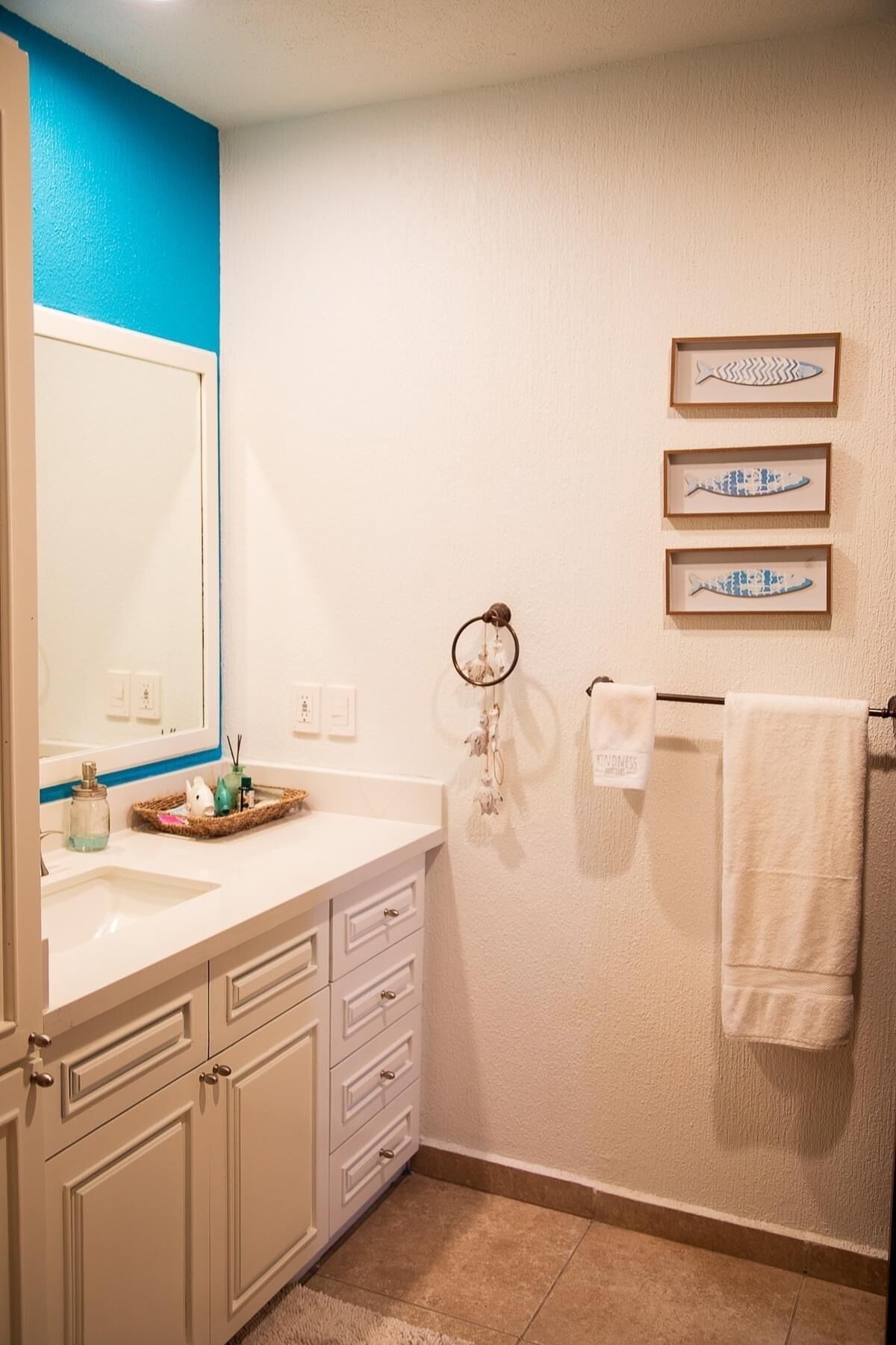 A bathroom with a white sink, beige cabinets, a blue accent wall, and framed fish artwork on the wall.