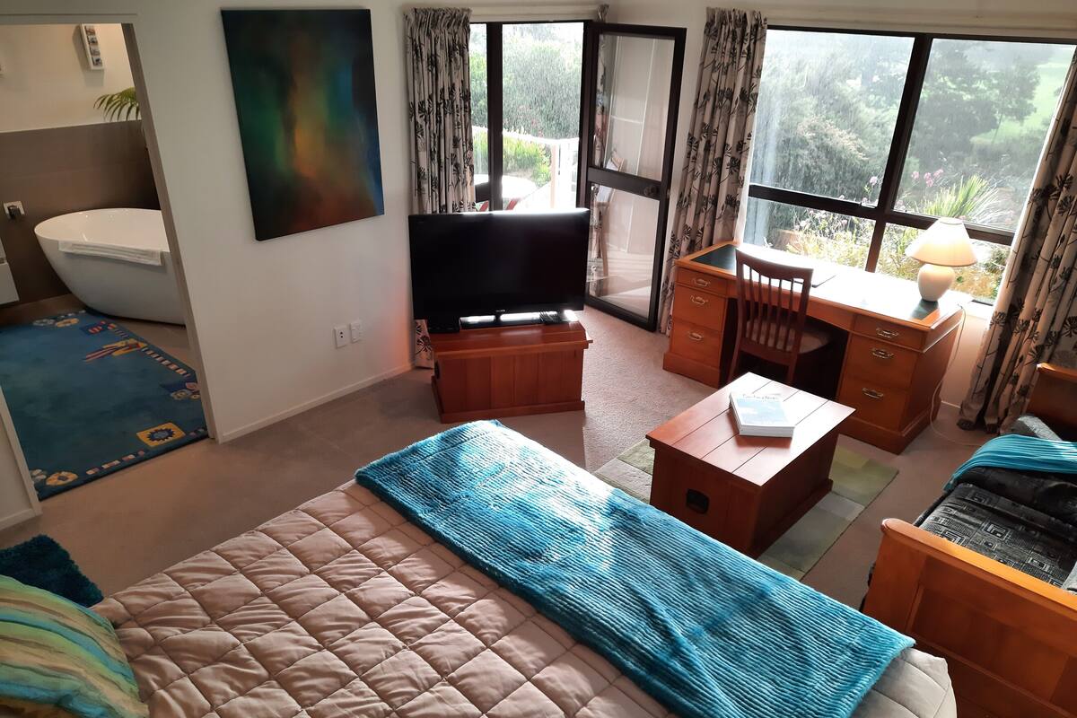 A bedroom with a bed, a desk, a television, and a view of a garden through the window.