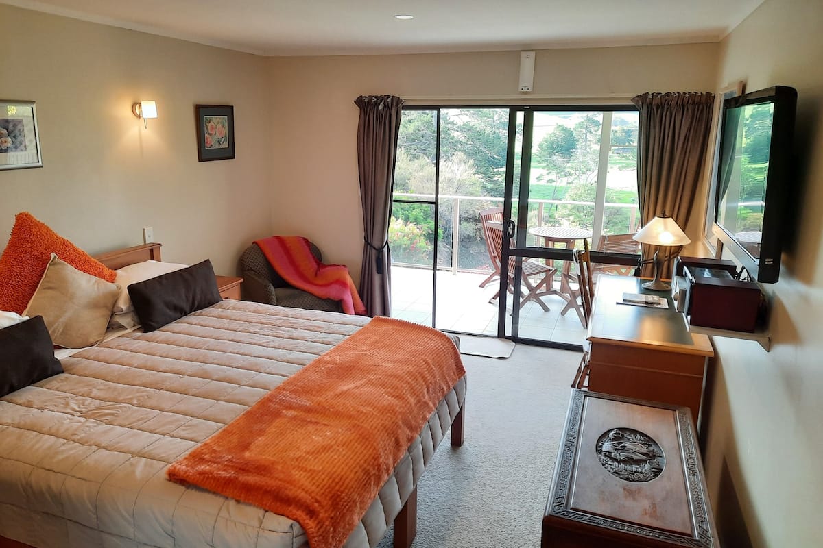 A hotel room with a bed, a desk with a lamp, a TV, and a balcony with a table and chairs.