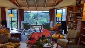 Un salon chaleureux avec vue sur un paysage verdoyant, une bibliothèque et un coin repas avec une table et des chaises.