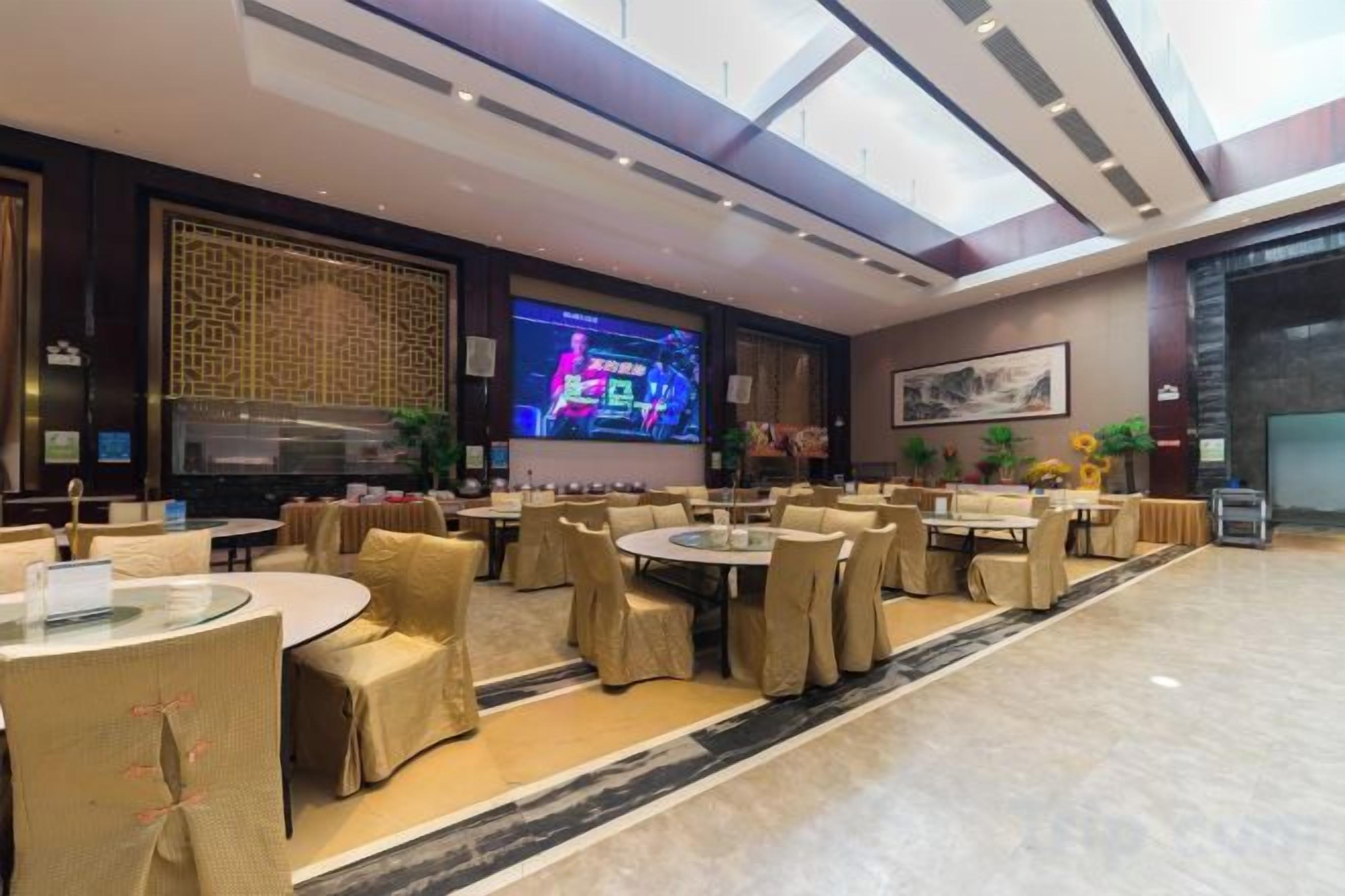 Un lobby espacioso de hotel con varias zonas de estar, una pantalla grande mostrando un programa y una decoración de interiores moderna.