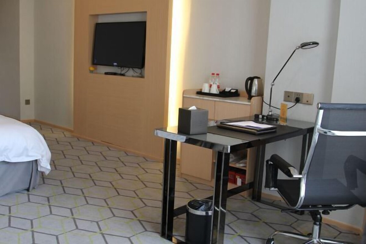 A hotel room with a bed, a desk with a chair, a television, a trash bin, and a small shelf with bottles and a kettle.