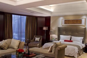 A hotel room with a large bed, two armchairs, a sofa, a coffee table with a vase of flowers, and a large window with curtains.