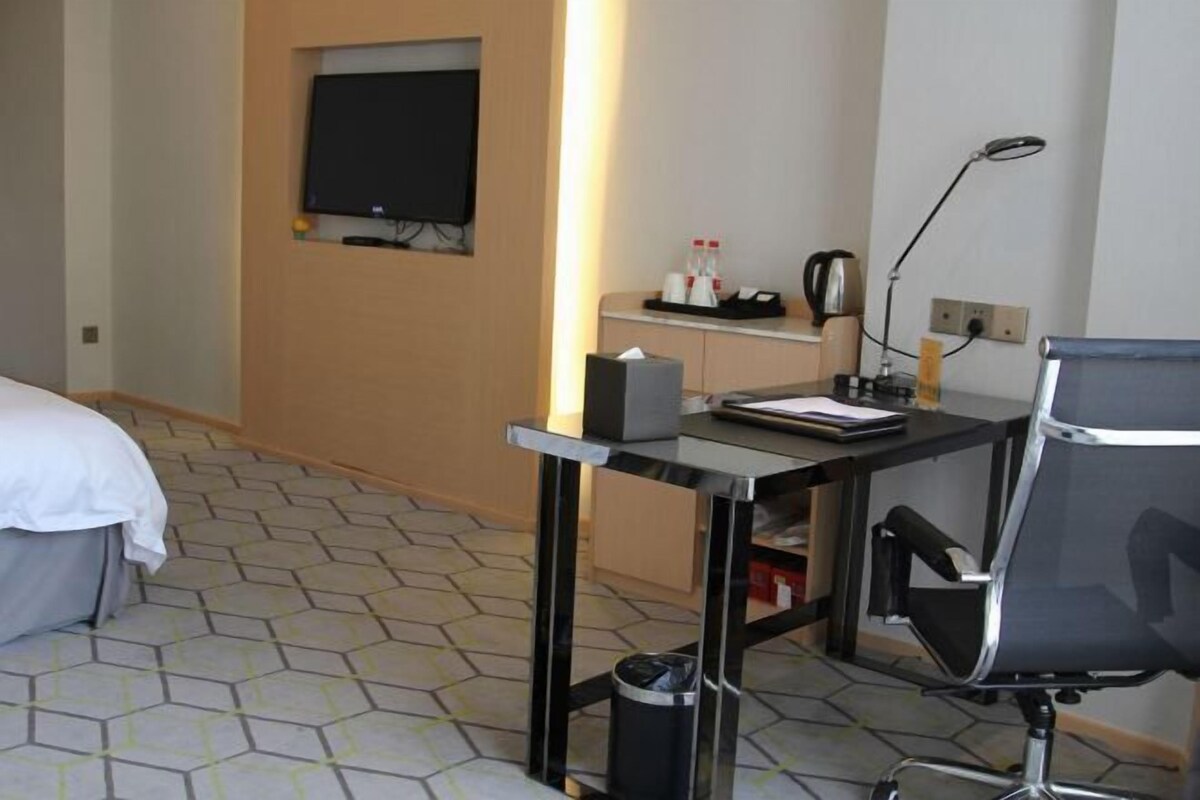A hotel room with a bed, a desk with a chair, a television, and a trash bin.