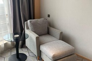 A corner room with a grey armchair, a glass table with a white object, and a window with curtains.